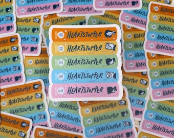 Op de afbeelding: Vijf kleurrijke stickers met de tekst "Heartstopper" en een volumenummer op elke sticker. De stickers zijn verticaal gerangschikt met de volumenummers in aflopende volgorde van boven naar beneden. De stickers zijn versierd met illustraties van twee personages uit de graphic novel.