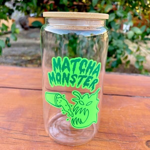 May include: A clear glass jar with a bamboo lid and a green and black sticker that says "MATCHA MONSTER". The sticker features a cartoon monster with green eyes and a mouth open wide, with green liquid pouring into it from a smaller cup.