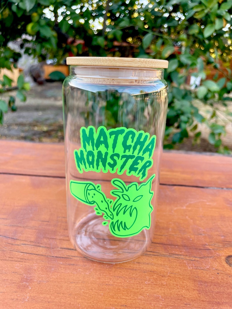 May include: A clear glass jar with a bamboo lid and a green and black sticker that says "MATCHA MONSTER". The sticker features a cartoon monster with green eyes and a mouth open wide, with green liquid pouring into it from a smaller cup.