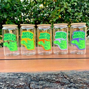 May include: Five clear glass jars with wooden lids and colorful labels that say "Matcha Monster". The labels feature a green monster with a green, orange, purple, yellow, and blue drink.