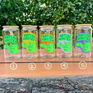 May include: Five clear glass jars with bamboo lids and green, orange, purple, and light green labels that say "Matcha Monster". The jars are lined up on a wooden surface.