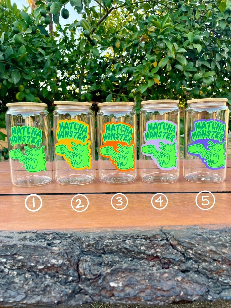 May include: Five clear glass jars with bamboo lids and green, orange, purple, and light green labels that say "Matcha Monster". The jars are lined up on a wooden surface.