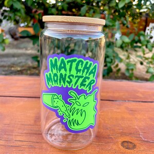 May include: A clear glass jar with a bamboo lid and a green and purple sticker that says "MATCHA MONSTER" with a cartoon monster drinking matcha.