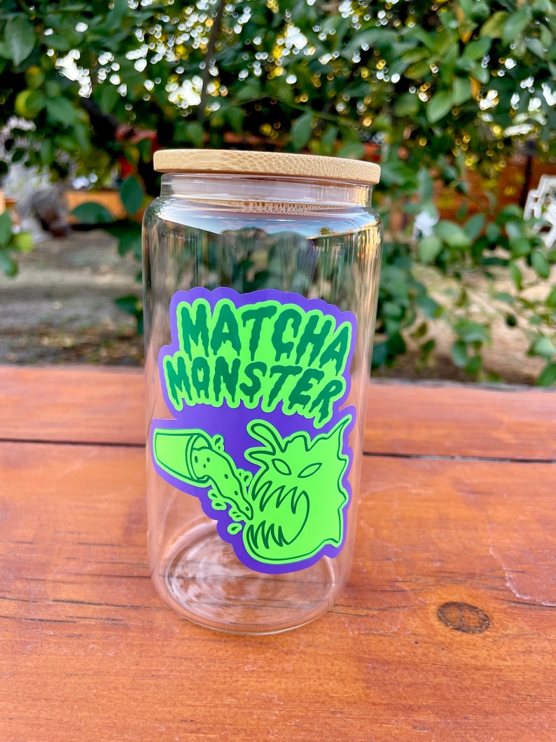 May include: A clear glass jar with a bamboo lid and a green and purple sticker that says "MATCHA MONSTER" with a cartoon monster drinking matcha.