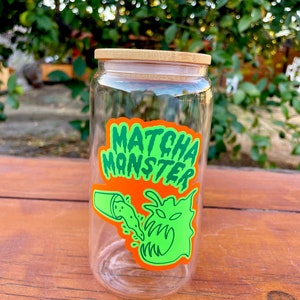 May include: A clear glass jar with a bamboo lid and a green and orange sticker that says "MATCHA MONSTER". The sticker features a cartoon monster with a mouth open and green liquid pouring into it.