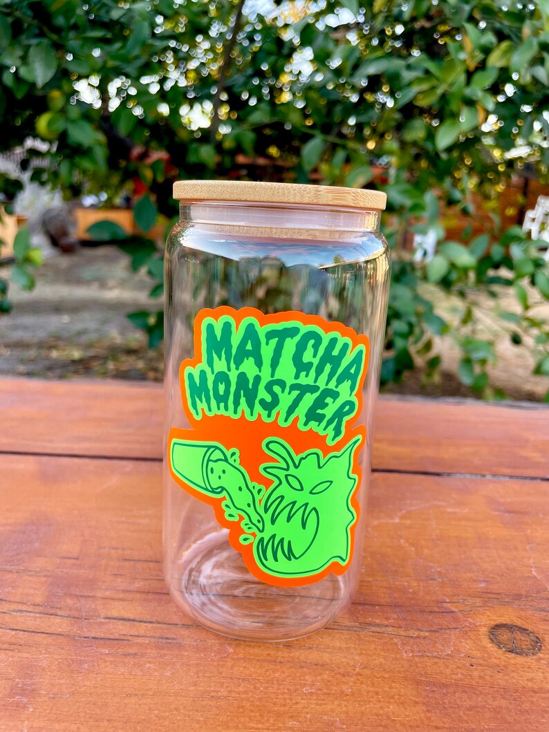 May include: A clear glass jar with a bamboo lid and a green and orange sticker that says "MATCHA MONSTER". The sticker features a cartoon monster with a mouth open and green liquid pouring into it.