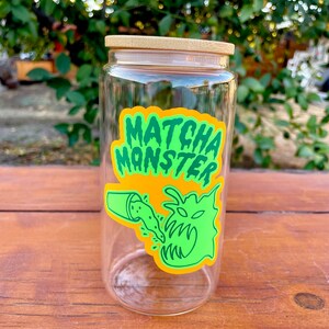 May include: A clear glass jar with a bamboo lid and a green and yellow sticker that says "Matcha Monster". The sticker features a cartoon monster with a mouth open and green liquid pouring into it.