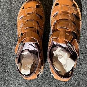 May include: A pair of brown leather sandals with a woven design. The sandals have a strap that goes around the ankle and a buckle closure.