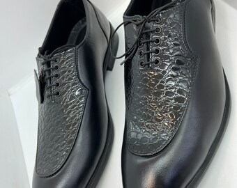 May include: A pair of black leather dress shoes. The shoes have a textured, crocodile-like pattern on the toe and vamp, with a smooth leather finish on the rest of the shoe. The shoes are laced up with black laces.