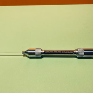 以下が含まれることがあります： 歯科用注射器のクローズアップ。注射器は金属製で、透明なガラス針が付いています。注射器にはリブ付きのグリップと端にノブが付いています。背景は明るい緑色です。