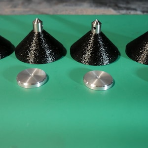 May include: Four black, cone-shaped objects with silver tips and four silver, circular discs are arranged on a green surface. The objects appear to be components of a larger item or set.