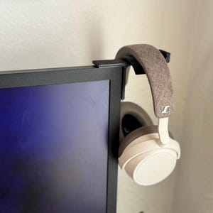 以下が含まれることがあります： コンピューターモニターに取り付けられた黒いヘッドホンハンガー。ベージュと茶色のヘッドホンが吊り下げられています。ヘッドホンにはゼンハイザーのロゴがあります。モニターには青い画面が表示されています。