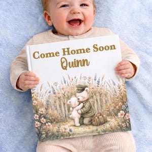 Può includere: Un libro quadrato con copertina rigida bianca e il testo dorato Come Home Soon Quinn. L'illustrazione mostra due orsacchiotti che si abbracciano in un campo di fiori selvatici vicino a una staccionata, uno indossa un berretto militare.