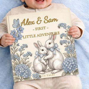 以下が含まれることがあります： 「Alex & Sam - First Little Adventure」というタイトルの子供向けの本。青いリボンをつけた2匹の白いウサギと、青い花のアクセントと蝶のイラストが描かれています。赤ちゃんが本を持っています。