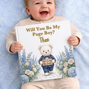 Może przedstawiać: Kwadratowa książka w twardej oprawie z białą okładką ze złotym napisem Will You Be My Page Boy? Theo. Ilustracja przedstawia pluszowego misia w niebieskim garniturze trzymającego tabliczkę z napisem Here Comes the Bride, otoczonego niebieskimi i białymi kwiatami.