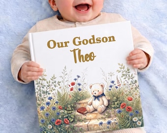 Puede incluir: Un libro cuadrado de tapa dura con el texto dorado Our Godson Theo en la portada blanca. La ilustración muestra un osito de peluche claro con un pañuelo azul sentado en escalones de piedra rodeado de flores silvestres y follaje verde.