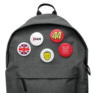 May include: A gray backpack with five round buttons attached. The buttons have various designs, including a British flag, a red and yellow racing car logo, a bulldog, the number 44, and the word "HAM".