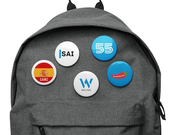 May include: A gray backpack with five round buttons attached. The buttons have various designs, including a Spanish flag with the word "SAINZ", a white button with the word "SAI", a blue button with the number "55", a white button with a "W" and the word "RACING", and a blue button with a red chili pepper and the text "Smooth Operator".