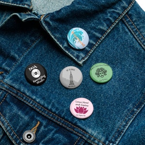 May include: Five different button pins on a blue denim jacket. The pins are round and feature various designs, including a record player, a building, a lotus flower, a mermaid, and a wave.