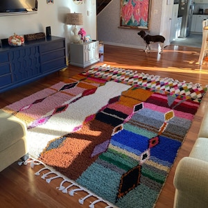 May include: A colorful, multi-patterned rug with fringe edges, laid out on a hardwood floor in a living room. The rug has a white center with pink, orange, blue, and green stripes and geometric shapes.