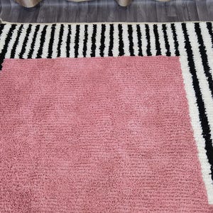 May include: A rectangular rug with a soft, textured pink center. The rug is bordered by a thick band of black and white vertical stripes. The rug is designed for home decor.