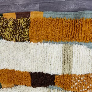 May include: A close-up of a colorful rug with a textured, geometric design. The rug features a mix of colors, including white, brown, yellow, and orange. The edges are finished with a green border and white tassels.
