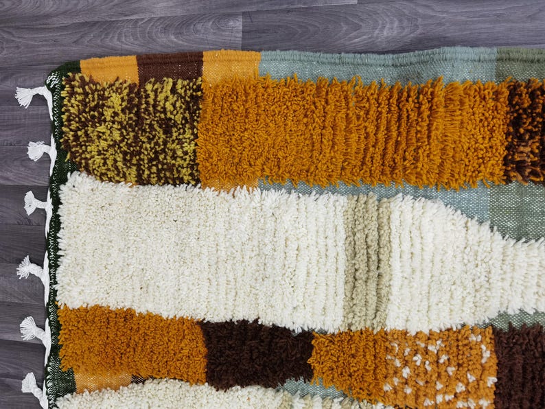 May include: A close-up of a colorful rug with a textured, geometric design. The rug features a mix of colors, including white, brown, yellow, and orange. The edges are finished with a green border and white tassels.