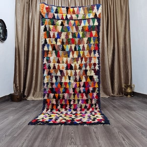 Puede incluir: Una alfombra marroquí colorida con un patrón geométrico de triángulos en tonos de rojo, azul, verde, amarillo y blanco. La alfombra está hecha de lana y tiene una textura tejida a mano.
