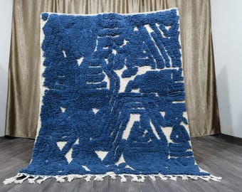 Può includere: Un tappeto rettangolare, fatto a mano, con un design geometrico blu navy e bianco. Il tappeto presenta un pelo spesso e strutturato e una frangia sul bordo inferiore. Il motivo include forme e linee astratte.
