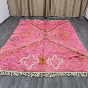 Op de afbeelding: Een roze Marokkaanse tapijt met een geometrisch patroon in tinten oranje, groen en wit. Het tapijt heeft een franje rand.