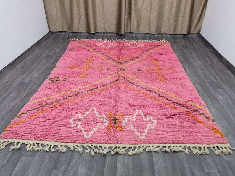 Op de afbeelding: Een roze Marokkaanse tapijt met een geometrisch patroon in tinten oranje, groen en wit. Het tapijt heeft een franje rand.