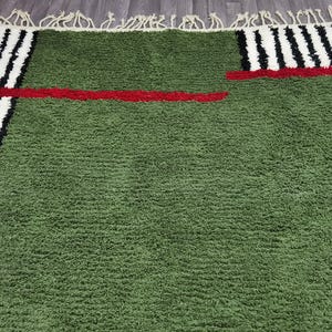 May include: A green rug with a textured surface. The rug features a red horizontal stripe and black and white vertical stripes on the edges. The rug has a fringe on one side.