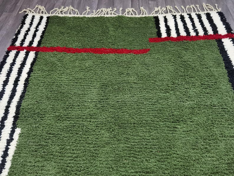 May include: A green rug with a textured surface. The rug features a red horizontal stripe and black and white vertical stripes on the edges. The rug has a fringe on one side.