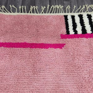 May include: A pink rug with a textured surface and a geometric design. The rug features a bright pink stripe and a section with black and white stripes. The top edge has a cream-colored fringe.