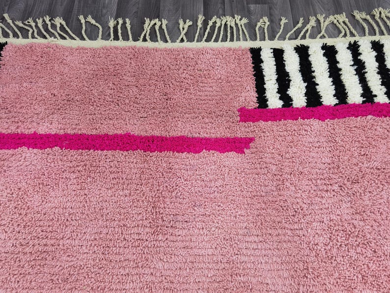May include: A pink rug with a textured surface and a geometric design. The rug features a bright pink stripe and a section with black and white stripes. The top edge has a cream-colored fringe.