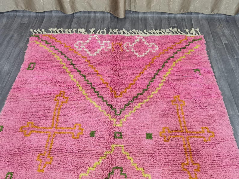 Op de afbeelding: Roze Marokkaanse tapijt met een geometrisch patroon met kruisen en zigzag in geel, oranje en groen.
