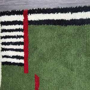 May include: A green rug with a textured surface. The rug features a design with black and white stripes, a red vertical stripe, and red accents. The rug has a fringe on one side.