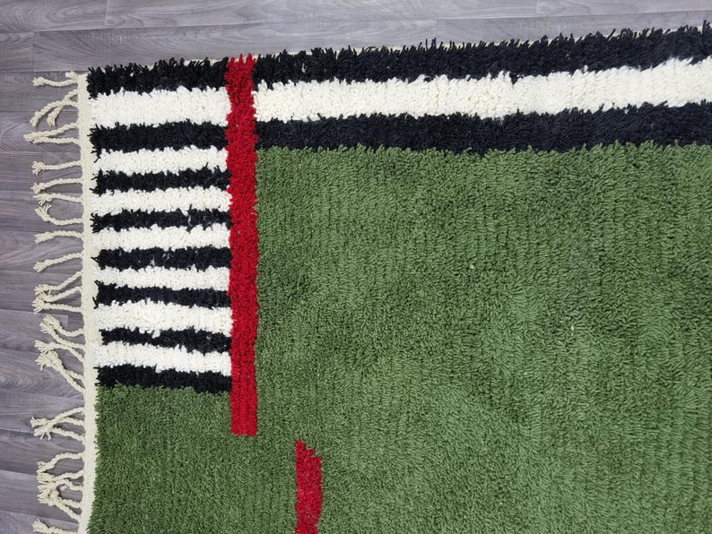 May include: A green rug with a textured surface. The rug features a design with black and white stripes, a red vertical stripe, and red accents. The rug has a fringe on one side.