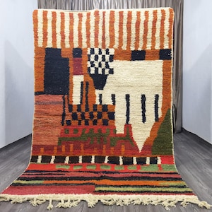 May include: A colorful Moroccan rug with geometric patterns in shades of orange, red, black, white, and green. The rug has a fringe border and is woven with a variety of textures.