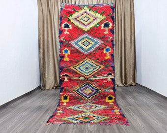 May include: A long, red Moroccan rug with a geometric pattern in shades of blue, yellow, and green. The rug has a fringe border and is woven with a thick, textured pile.