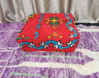 May include: A red, square pouf with geometric patterns in orange, green, yellow, and blue. The pouf is placed on a purple and white patterned rug, with a gray curtain in the background. The pouf is a decorative piece.