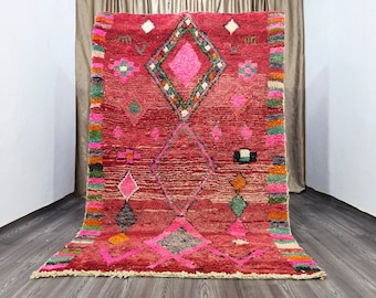 May include: A pink and red Moroccan rug with a diamond pattern and colorful accents. The rug has a fringe border and is made of wool.