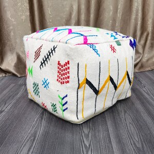 K&ouml;nnte beinhalten: Ein wei&szlig;er quadratischer Hocker mit bunten geometrischen Mustern, die in den Stoff eingewebt sind. Der Hocker steht auf einem Holzboden.