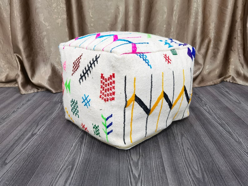 K&ouml;nnte beinhalten: Ein wei&szlig;er quadratischer Hocker mit bunten geometrischen Mustern, die in den Stoff eingewebt sind. Der Hocker steht auf einem Holzboden.