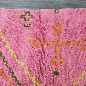 Op de afbeelding: Roze kleed met een geometrisch patroon in geel, oranje en groen. Het patroon bevat een groot kruisvormig figuur in het midden.