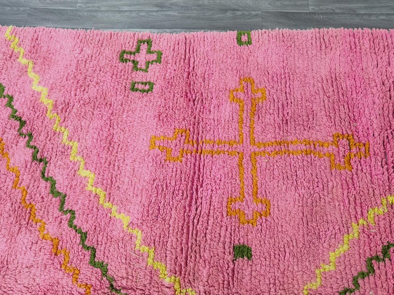 Op de afbeelding: Roze kleed met een geometrisch patroon in geel, oranje en groen. Het patroon bevat een groot kruisvormig figuur in het midden.