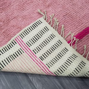 May include: A pink rug with a textured surface and a geometric design. The underside of the rug is visible, featuring a cream-colored background with black and pink stripes, and fringe detailing.