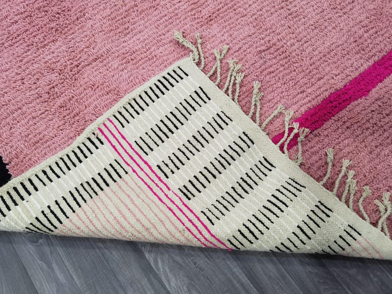 May include: A pink rug with a textured surface and a geometric design. The underside of the rug is visible, featuring a cream-colored background with black and pink stripes, and fringe detailing.