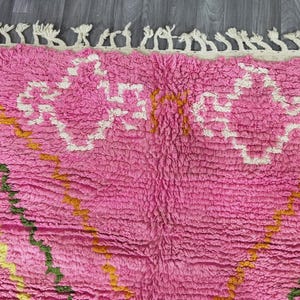 Op de afbeelding: Roze en wit Marokkaans tapijt met een geometrisch patroon. Het tapijt heeft een franje rand en is gemaakt van een zacht, pluche materiaal.