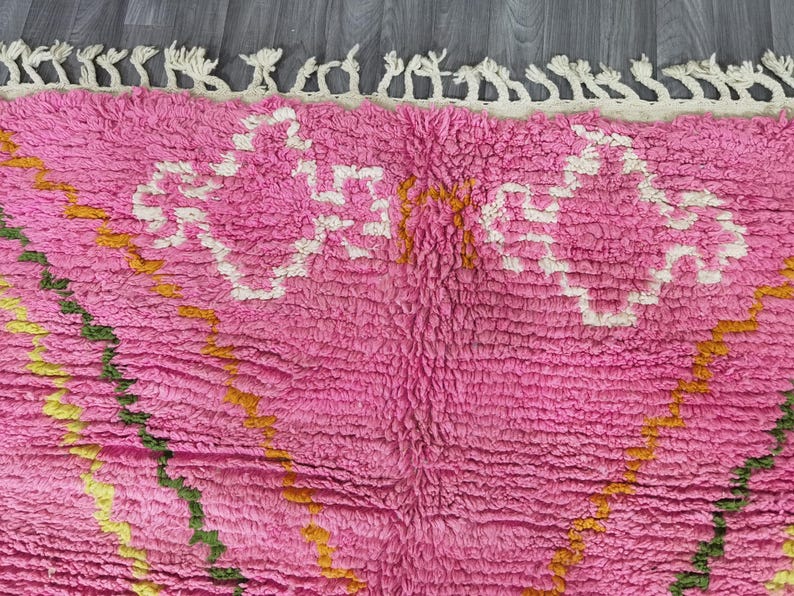 Op de afbeelding: Roze en wit Marokkaans tapijt met een geometrisch patroon. Het tapijt heeft een franje rand en is gemaakt van een zacht, pluche materiaal.